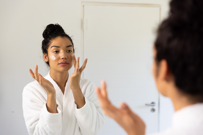 Biracial woman in bathrobe applying cream on her face in sunny bathroom. Lifestyle, self care, beauty, skin and domestic life, unaltered.